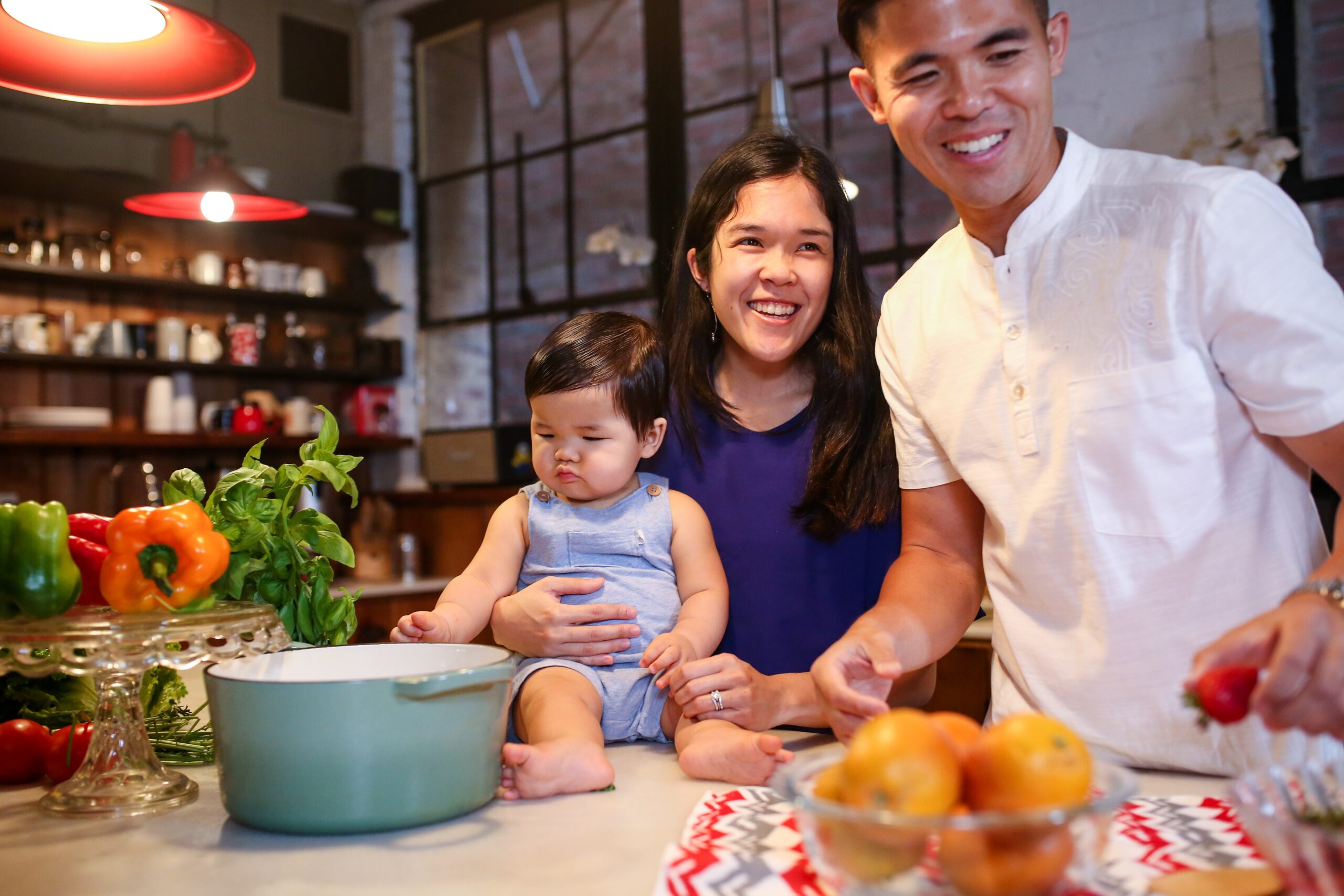 Young couple in their kitchen with dad cooking and mom holding cute baby on the counter