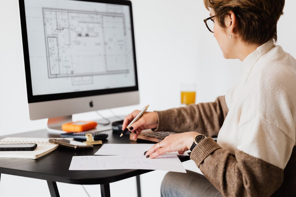 Middle aged woman sitting at desk working on a computer with architectural drawings on the screen. 
